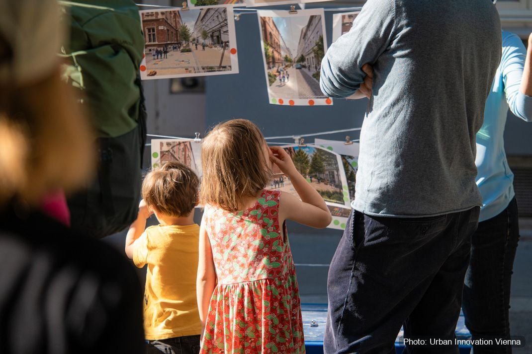 Children and adults in the street looking up at printed scenarios pegged to a line.
