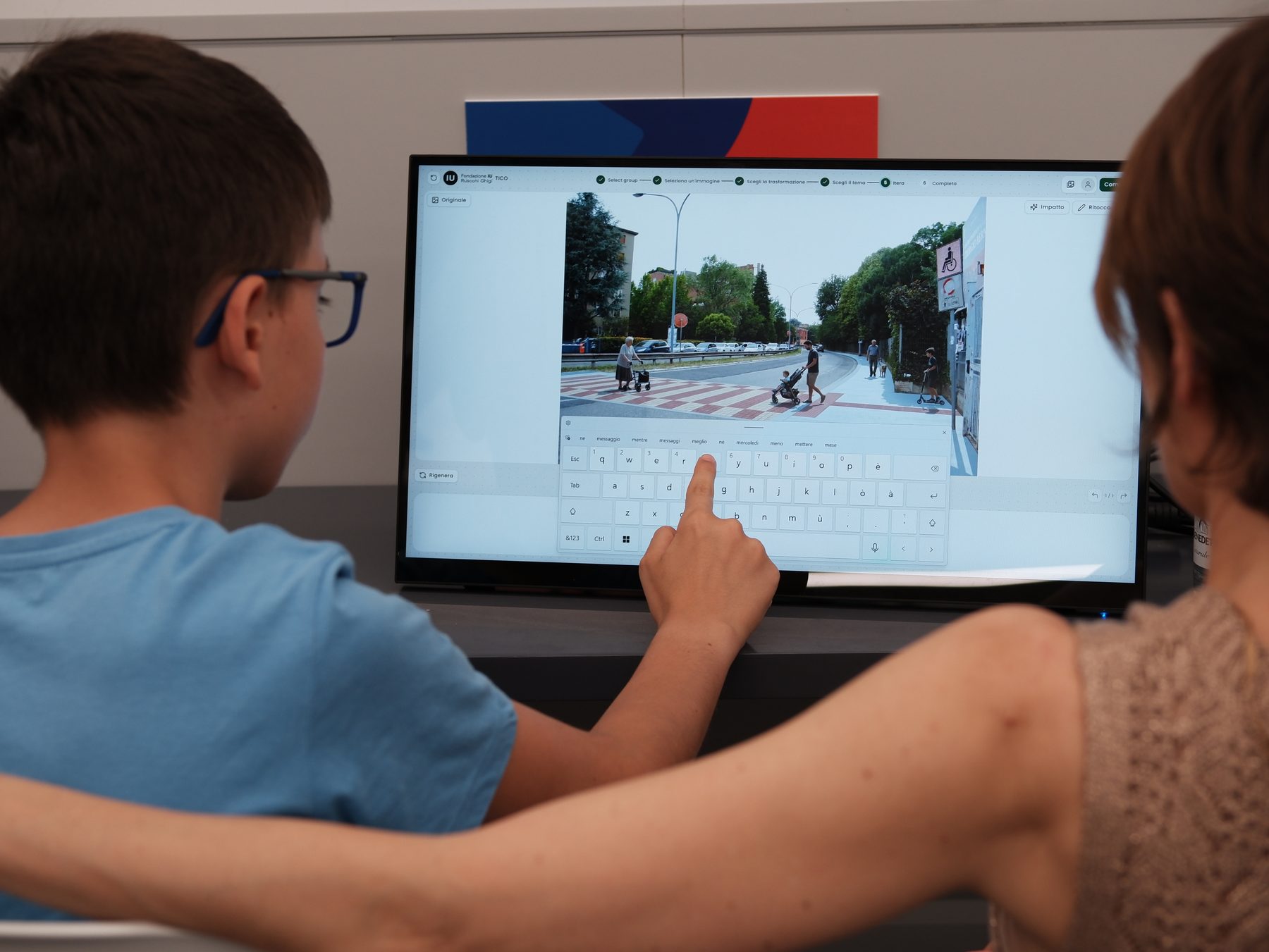 A boy types on a large touchscreen that shows a photograph of a tree-lined street, an adult’s arm resting on his shoulder.