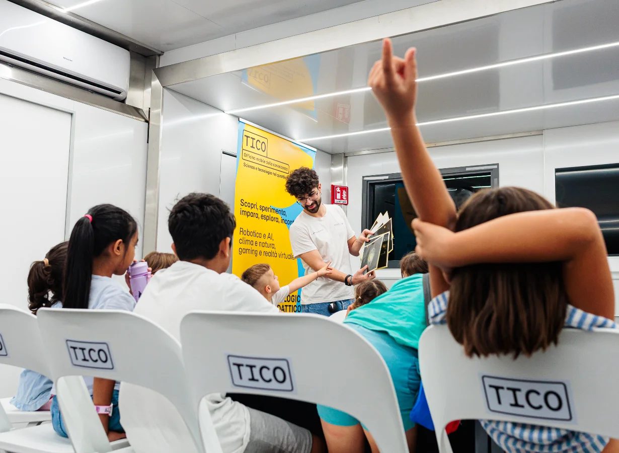 Children on chairs inside the TICO truck, one hand raised, a screen at the front.